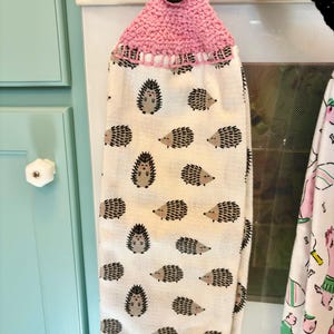 Op de afbeelding: Een hangende keukendoek met een herhalend patroon van kleine bruine en zwarte egels op een cr&egrave;mekleurige achtergrond. Het bovenste gedeelte is gemaakt van roze gehaakt garen en heeft een enkele donkerbruine knoop om de doek vast te zetten.