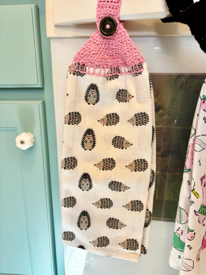 Op de afbeelding: Een hangende keukendoek met een herhalend patroon van kleine bruine en zwarte egels op een cr&egrave;mekleurige achtergrond. Het bovenste gedeelte is gemaakt van roze gehaakt garen en heeft een enkele donkerbruine knoop om de doek vast te zetten.