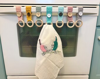 Puede incluir: Conjunto de siete agarraderas para manoplas de horno de ganchillo con anillos de madera. Cada agarradera tiene un color diferente y un botón. Las agarraderas están colgadas en una puerta de horno blanca.
