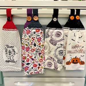 May include: A collection of Halloween-themed kitchen towels with crochet tops and button closures. Designs include skeletons, floral patterns, crystal balls, and pumpkins. One towel reads "Bone appetite!" and another says "Happy Halloween".