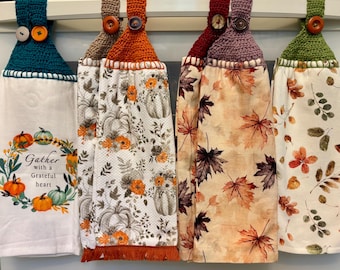 Puede incluir: Una colección de cinco paños de cocina colgantes con la parte superior de ganchillo y cierres de botón. Las toallas presentan diseños con temas de otoño, incluyendo calabazas, hojas y diseños florales. Una toalla tiene el texto "Gather with a Grateful Heart."