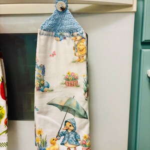 Op de afbeelding: Een hangende keukendoek van witte stof met een kleurrijke print van eendjes in regenkleding, paraplu's en lentebloemen. De doek is aan de bovenkant voorzien van een lichtblauw gehaakt lusje dat met een witte knoop wordt vastgezet.