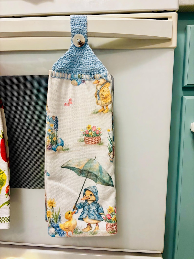 Op de afbeelding: Een hangende keukendoek van witte stof met een kleurrijke print van eendjes in regenkleding, paraplu's en lentebloemen. De doek is aan de bovenkant voorzien van een lichtblauw gehaakt lusje dat met een witte knoop wordt vastgezet.
