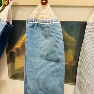Op de afbeelding: Een lichtblauwe theedoek met wafelstructuur, voorzien van een wit gehaakt bovendeel met een ophanglus en een bruine knoop. De handdoek is gemaakt van gestructureerde stof en is met witte stiksels aan het gehaakte bovendeel bevestigd.