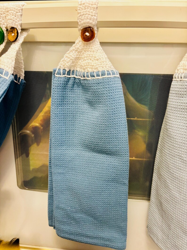 Op de afbeelding: Een lichtblauwe theedoek met wafelstructuur, voorzien van een wit gehaakt bovendeel met een ophanglus en een bruine knoop. De handdoek is gemaakt van gestructureerde stof en is met witte stiksels aan het gehaakte bovendeel bevestigd.