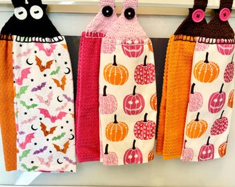 Puede incluir: Tres toallas de cocina con temática de Halloween con lazos de colgar de ganchillo. Las toallas presentan una combinación de colores rosa, naranja y negro. Una toalla tiene un patrón de murciélago, otra tiene un patrón de calabaza y otra tiene un patrón de calabaza y fantasma.