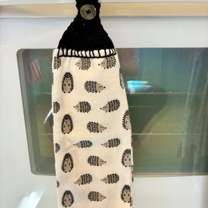 Op de afbeelding: Een hangende keukendoek met een herhalend patroon van kleine bruine en zwarte egels op een cr&egrave;mekleurige achtergrond. Het bovenste gedeelte is gemaakt van zwart gehaakt garen en bevat een enkele ronde knoop om de doek vast te zetten.