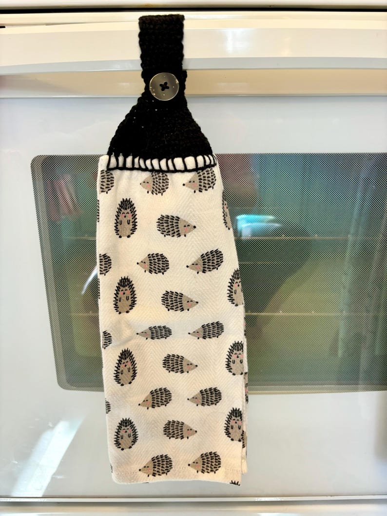 Op de afbeelding: Een hangende keukendoek met een herhalend patroon van kleine bruine en zwarte egels op een cr&egrave;mekleurige achtergrond. Het bovenste gedeelte is gemaakt van zwart gehaakt garen en bevat een enkele ronde knoop om de doek vast te zetten.