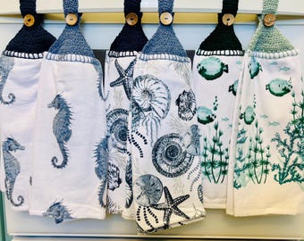 Op de afbeelding: Zes hangende keukendoeken met gehaakte bovenkanten in marineblauw, blauw en saliegroen, elk bevestigd met een houten knoop. De witte stof is voorzien van diverse zeeprints zoals zeepaardjes, schelpen, zeesterren en waterplanten.
