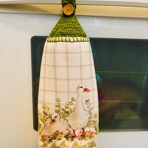 Op de afbeelding: Een hangende keukendoek van witte stof met een lichtbeige ruitpatroon en een aquarelillustratie van twee witte ganzen tussen groen gebladerte en rode bessen. De bovenkant heeft een groene gehaakte hanger met een ronde houten knoop.