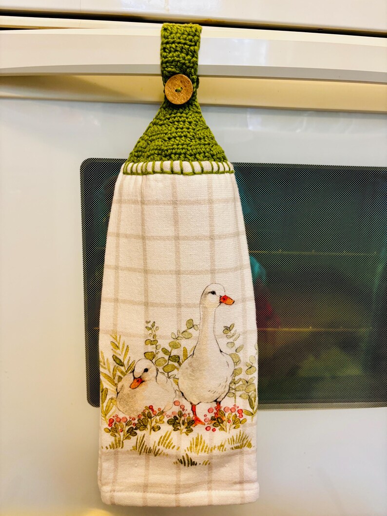 Op de afbeelding: Een hangende keukendoek van witte stof met een lichtbeige ruitpatroon en een aquarelillustratie van twee witte ganzen tussen groen gebladerte en rode bessen. De bovenkant heeft een groene gehaakte hanger met een ronde houten knoop.