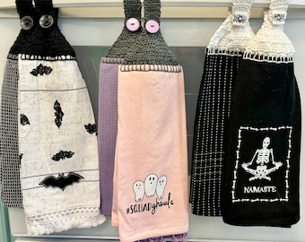 Puede incluir: Tres toallas de cocina con temática de Halloween colgadas en un estante. La toalla de la izquierda tiene un patrón de murciélago blanco y negro. La toalla del medio es rosa con un diseño de fantasma y el texto "#squadghouls". La toalla de la derecha es negra con un esqueleto en una pose de yoga y el texto "NAMASTE".