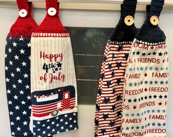 Puede incluir: Dos juegos de toallas de cocina rojo, blanco y azul con lazos de ganchillo. Un juego presenta una caravana rojo, blanco y azul con el texto "Happy 4th of July". El otro juego presenta el texto "Freedom Friends Family" repetido en un patrón.