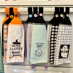 May include: A collection of Halloween-themed kitchen towels hanging on a metal bar. The towels feature crochet tops with button closures and various designs, including a witch hat, the phrase "Just a Little Hocus Pocus," and a ghost with the words "Did someone say BOOS?"