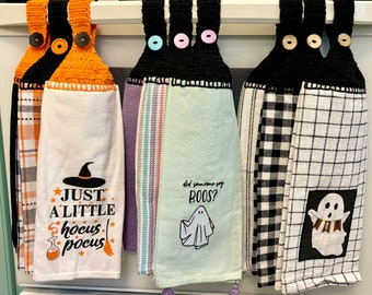 Puede incluir: Una colección de paños de cocina con temática de Halloween colgados de una barra metálica. Los paños presentan la parte superior de ganchillo con cierres de botón y varios diseños, incluyendo un sombrero de bruja, la frase "Just a Little Hocus Pocus" y un fantasma con "Did someone say BOOS?"