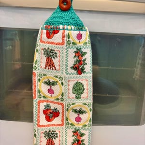 Op de afbeelding: Een hangende keukendoek van witte stof met een ruitpatroon van kleurrijke groente-illustraties, waaronder wortels, tomaten, radijzen en artisjokken. De bovenkant heeft een turquoise gehaakte hanger die vastzit met een bruine knoop.