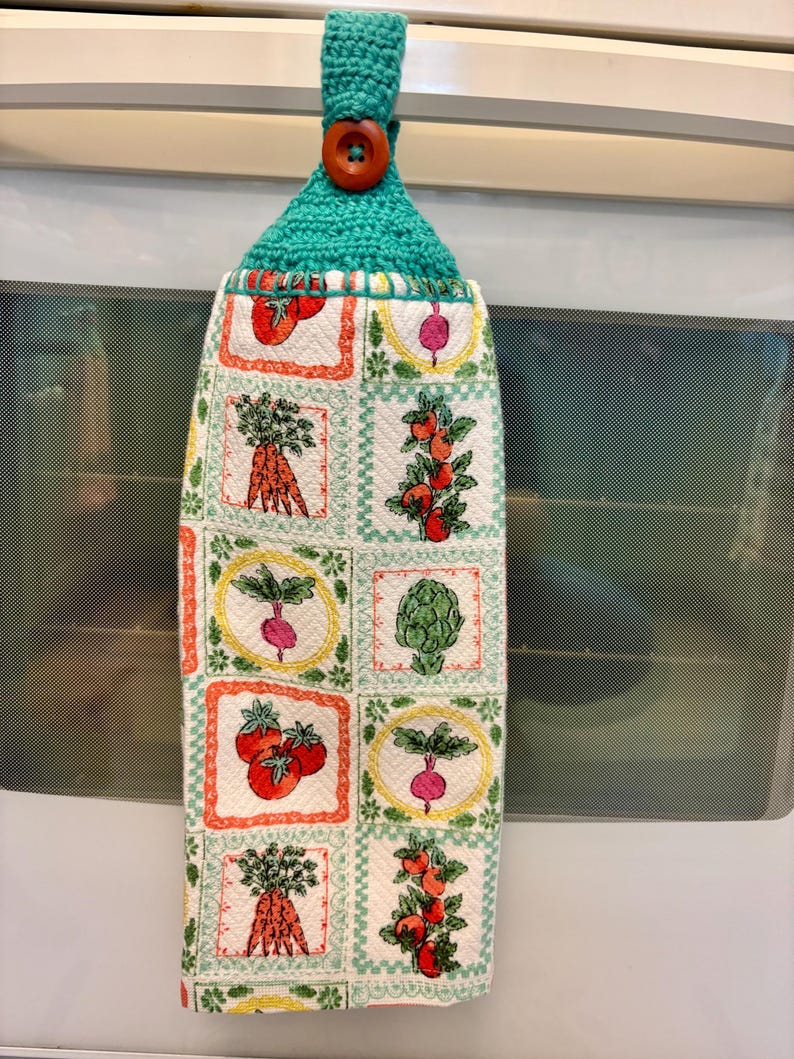 Op de afbeelding: Een hangende keukendoek van witte stof met een ruitpatroon van kleurrijke groente-illustraties, waaronder wortels, tomaten, radijzen en artisjokken. De bovenkant heeft een turquoise gehaakte hanger die vastzit met een bruine knoop.
