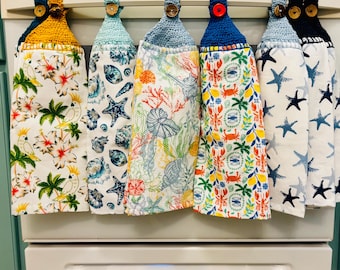 Puede incluir: Una fila de siete paños de cocina colgantes con partes superiores de ganchillo en varios colores, cada uno asegurado con un botón. Los paños presentan diversos patrones que incluyen aves tropicales, conchas marinas, arrecifes de coral, langostas y estrellas de mar.