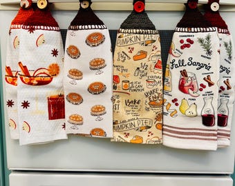 Puede incluir: Seis paños de cocina colgantes con partes superiores de ganchillo y botones, con varios estampados de temática otoñal que incluyen pasteles, recetas de especias de calabaza e ingredientes de sangría con texto sobre tela blanca y crema.