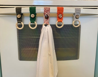 Op de afbeelding: Vijf gehaakte handdoekringen met houten ringen en knopen die aan een witte ovendeur hangen. De ringen zijn in verschillende kleuren, waaronder groen, rood, oranje en grijs.