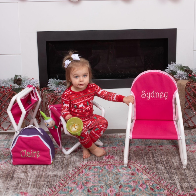 Puede incluir: Dos sillas de ni&ntilde;os rosas con marcos blancos. Una silla tiene el nombre "Sydney" bordado en la parte posterior y la otra silla tiene el nombre "Claire" bordado en la parte delantera. Una ni&ntilde;a peque&ntilde;a en pijama rojo y blanco est&aacute; sentada en una de las sillas.