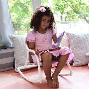 Puede incluir: Una ni&ntilde;a peque&ntilde;a con un vestido rosa con el nombre "Lily" bordado en &eacute;l, se sienta en una silla mecedora blanca y lee un libro. Lleva una flor rosa en el pelo. La silla est&aacute; sobre un suelo de baldosas rosa y blanco.