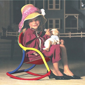 Puede incluir: Una ni&ntilde;a peque&ntilde;a con un sombrero de paja rosa se sienta en una silla mecedora colorida. Est&aacute; sosteniendo una mu&ntilde;eca y la est&aacute; alimentando con un biberon. La silla est&aacute; hecha de pl&aacute;stico rojo, amarillo, azul y verde.