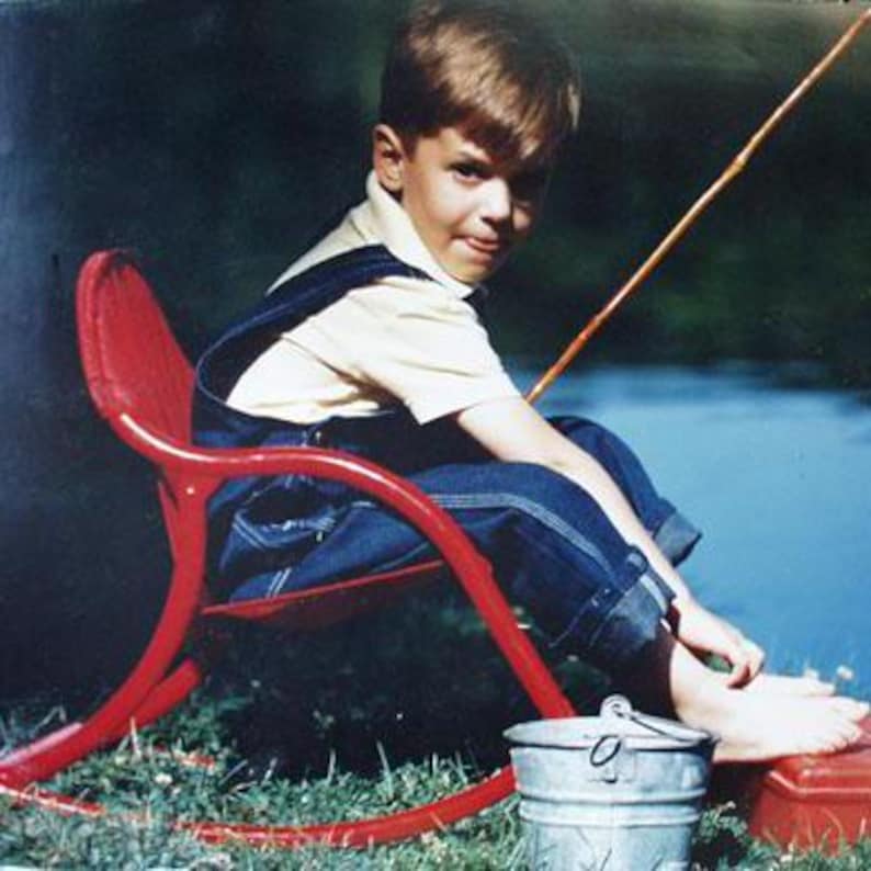 Pode incluir: Um menino pequeno est&aacute; sentado em uma cadeira de balan&ccedil;o vermelha com uma vara de pesca na m&atilde;o. Ele est&aacute; vestindo jeans azul e uma camisa amarela. A cadeira est&aacute; em uma superf&iacute;cie gramada.