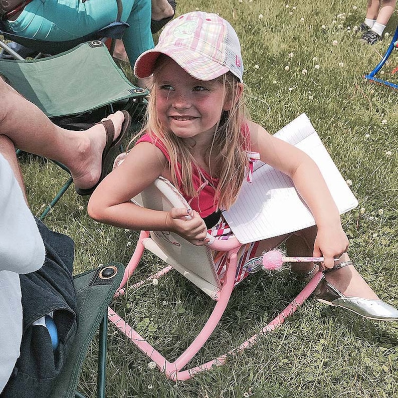 Puede incluir: Una ni&ntilde;a peque&ntilde;a con una gorra de b&eacute;isbol de cuadros rosa y blanco se sienta en una silla mecedora rosa. Ella sostiene un cuaderno y un bol&iacute;grafo. La silla est&aacute; en un &aacute;rea cubierta de hierba.