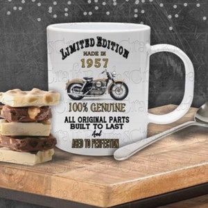 Op de afbeelding: Witte keramische mok met een motorfietsafbeelding en de tekst "LIMITED EDITION MADE IN 1957". De mok heeft ook de tekst "100% GENUINE ALL ORIGINAL PARTS BUILT TO LAST AND AGED TO PERFECTION". Een zilveren lepel en een stapel chocolade liggen op een houten oppervlak.