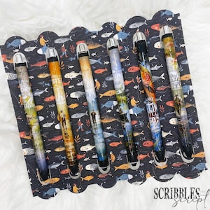 May include: Set of six black pens with colorful designs featuring fish and fishing-related text. The pens have silver caps and are arranged on a black background with a white scalloped border. The text on the pens includes "Reel a Perfect Catch", "Lake Life", "Keep Fishin' Great", "I'd Rather Be Fishin'" and "The Lure is Real".