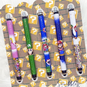 May include: Four pens with different Super Mario Bros. characters on them. The pens are displayed in a cardboard holder with a question mark pattern. The text "Scribbles & Script" is at the bottom of the holder.