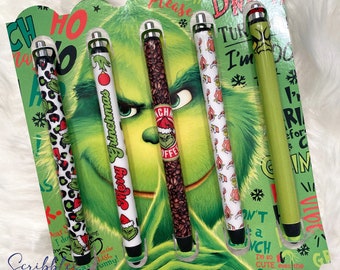 Peut inclure: Un ensemble de six stylos sur le thème du Grinch avec différents motifs, dont un imprimé léopard, une tasse à café et un Grinch disant "Joyeux Grinchmas". Les stylos sont emballés dans une carte verte et blanche avec une illustration du Grinch.