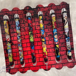 May include: Six black pens with silver caps are displayed on a red brick background. Each pen features a different comic book character, including Spider-Man, Captain America, and Iron Man.
