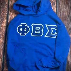 Könnte beinhalten: Ein königsblauer Kapuzenpullover mit weißen und marinefarbenen griechischen Buchstaben. Die Buchstaben sind groß und mittig auf der Vorderseite. Der Hoodie hat eine Kapuze mit Kordelzug und eine Vordertasche.