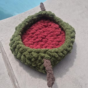 May include: A handmade watermelon-shaped pet bed. The bed features a plush, textured design with a red interior, a green rind, and a brown stem. The bed is designed to resemble a slice of watermelon.