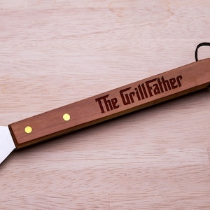 May include: A silver spatula with a wooden handle. The handle has the words "The GrillFather" engraved on it.