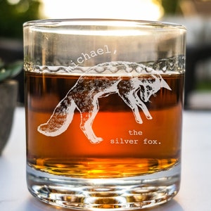 May include: A clear glass tumbler with a brown liquid inside. The glass has a design etched into it of a fox jumping with the text "Michael, the silver fox."