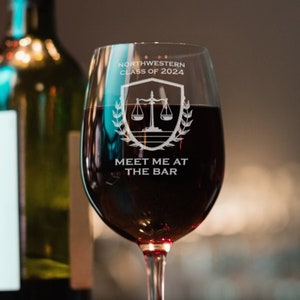 Puede incluir: Una copa de vino transparente con un diseño blanco que dice "Northwestern Class of 2024" y "Meet me at the bar". El diseño también incluye una balanza de la justicia y una corona de laurel. La copa está llena de vino tinto y está sobre una mesa de madera.