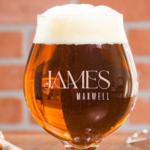 Può includere: Un bicchiere di birra personalizzato con il nome "JAMES MAXWELL" inciso sul lato. Il bicchiere è pieno di una birra ambrata scura e ha uno spesso strato di schiuma in cima.