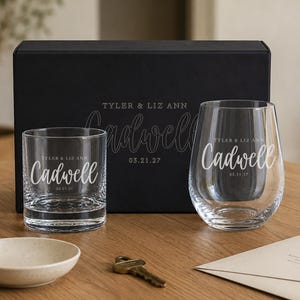 May include: A black rectangular gift box featuring the text Tyler & Liz Ann Cadwell 03.21.27, accompanied by a short rocks glass and a stemless wine glass, both etched with the same names and date. A small beige bowl, a key, and an envelope are present.