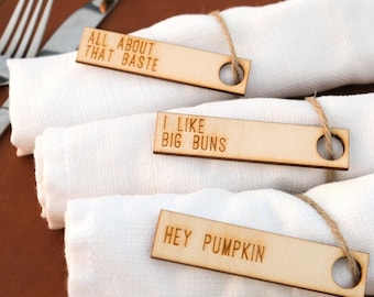 May include: Wooden napkin rings with engraved text: "ALL ABOUT THAT BASTE", "I LIKE BIG BUNS", and "HEY PUMPKIN". The rectangular tags are attached to white cloth napkins with twine. The setting includes a brown placemat and silverware.