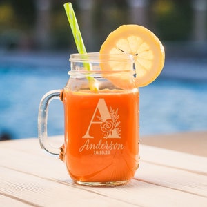 Puede incluir: Un mug de vidrio de tipo mason jar personalizado con una pajita, lleno de zumo de naranja y una rodaja de limón. El mug está grabado con la letra "A", el nombre "Anderson" y la fecha "10.10.20".
