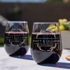 Eye-level view of two engraved stemless wine glasses. The etching has a circular leafy border with the names Matt & Lilliana in the center with Est. 10.25.22 underneath them. The glasses are sitting on a white outdoor table.