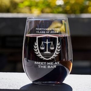 May include: A clear stemless wine glass with a design etched on the side. The design features a scale of justice, a laurel wreath, and the text "Northwestern Class of 2024 Meet Me At The Bar". The glass is filled with red wine.
