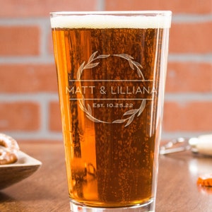 May include: A frosted glass beer mug with a personalized design. The design features a wreath with leaves and the text "Matt & Lilliana Est. 10.25.22".