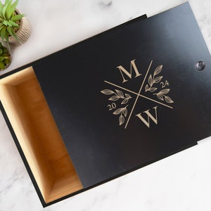 May include: A black wooden box with a hinged lid. The lid is engraved with a monogram of the letters "M" and "W" with a floral design and the dates "20" and "24".