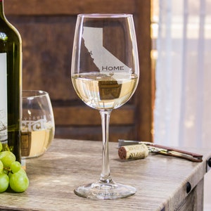 May include: A clear glass wine glass with a white wine inside. The glass is etched with the outline of the state of California and the word "HOME".
