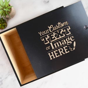 May include: Black wooden box with a hinged lid, featuring the text "Your Custom TEXT or Image HERE" in a light brown color. The box is partially open, revealing a light brown interior. A small plant is visible in the background.