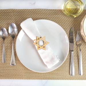 May include: A white dinner plate with a white napkin and a wooden Star of David napkin ring on a brown placemat. The plate is set with a fork, knife, and spoon.